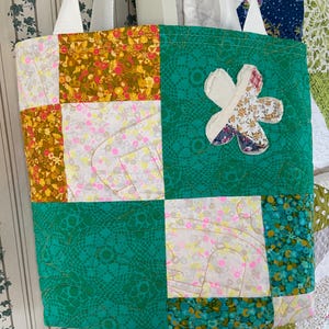 May include: A quilted tote bag with a patchwork design. The bag features a large green square with a flower applique. The patchwork includes floral and patterned fabrics in shades of yellow, pink, and teal. White straps complete the design.