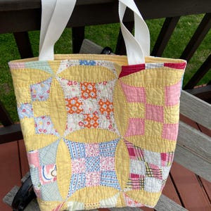 May include: A handmade tote bag with a patchwork design. The bag features a yellow background with colorful fabric squares in pink, blue, orange, and white. It has white handles and a red trim.