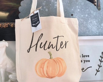 Op de afbeelding: Een witte canvas draagtas met de naam "Hunter" gedrukt in zwarte schrijfletters. Een kleine oranje pompoen met een groene steel is op de tas gedrukt. De tas staat op een houten tafel met een wit fotolijstje en een kleine plant in een pot.