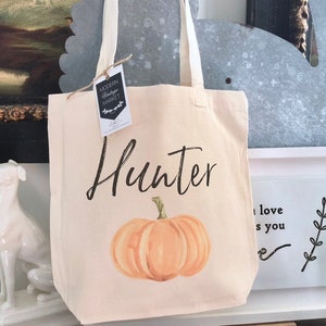 Może przedstawiać: Biała torba z płótna z nadrukowanym imieniem "Hunter" czarnym pismem. Na torbie nadrukowana jest mała pomarańczowa dynia z zieloną łodygą. Torba stoi na drewnianym stole z białą ramką i małą rośliną w doniczce.