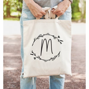 May include: A white canvas tote bag with black floral wreath and the letter "M" in the center. The bag is being held by a person wearing a gray shirt and blue jeans.