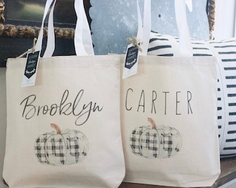 Puede incluir: Dos bolsas de lona blancas con asas negras. Las bolsas tienen un diseño de calabaza a cuadros en blanco y negro y los nombres "Brooklyn" y "Carter" impresos en ellas.
