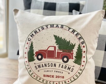 May include: A white pillow with a red truck carrying a Christmas tree. The truck is surrounded by a green circle with the text "Christmas Trees", "Swanson Farms", "Family Owned", "Since 2018", "Pine", "Spruce", "Fir", and "Cedar".