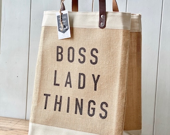 Puede incluir: Un bolso tote rectangular de yute beige con ribetes color crema. Las palabras "BOSS LADY THINGS" están impresas en letras negras grandes. Tiene asas de cuero marrón y una etiqueta.