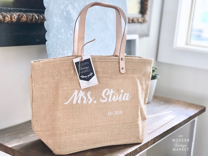 Op de afbeelding: Een natuurlijke jute tas met leren handvatten en een witte inscriptie met de tekst "Mrs. Stoia Est. 2019". Een klein label met de tekst "MODERN Vintage MARKET" is aan de tas bevestigd.