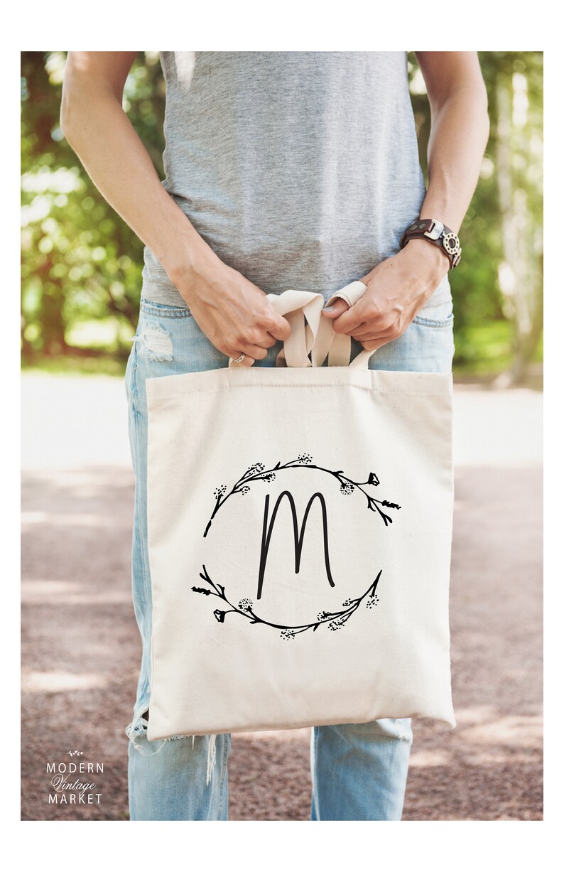 May include: A white canvas tote bag with black floral wreath and the letter "M" in the center. The bag is being held by a person wearing a gray shirt and blue jeans.
