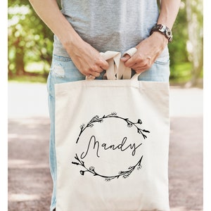 May include: A white canvas tote bag with black floral wreath and the name "Mandy" printed in a cursive font.