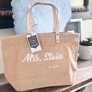 Op de afbeelding: Een natuurlijke jute draagtas met leren handvatten en een witte inscriptie met de tekst "Mrs. Stoia Est. 2019".