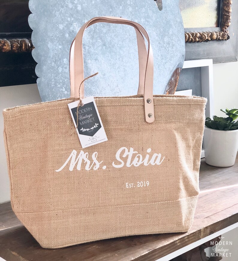 Op de afbeelding: Een natuurlijke jute draagtas met leren handvatten en een witte inscriptie met de tekst "Mrs. Stoia Est. 2019".