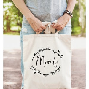 May include: A white canvas tote bag with black floral wreath and the name "Mandy" printed on it.