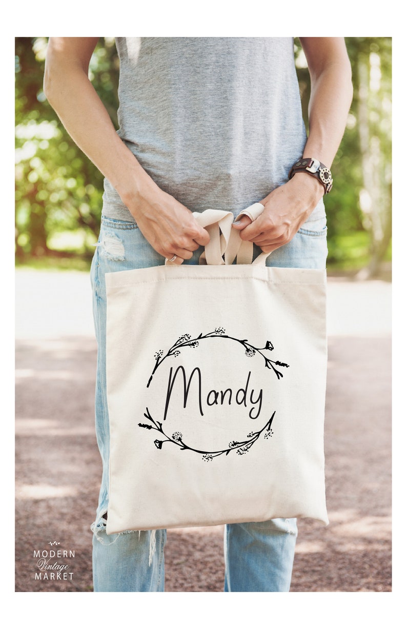 May include: A white canvas tote bag with black floral wreath and the name "Mandy" printed on it.