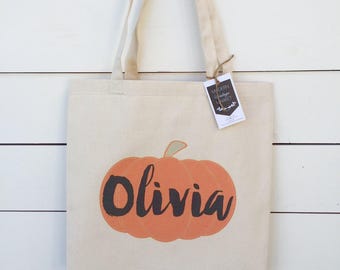 Op de afbeelding: Een witte canvas draagtas met een pompoenontwerp en de naam "Olivia" in zwart gedrukt. De tas heeft een bruin label met de tekst "Modern Vintage Market" erop.