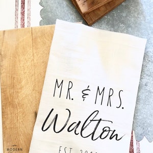 May include: White kitchen towel with black text that reads "MR. & MRS. Walton EST. 2018".