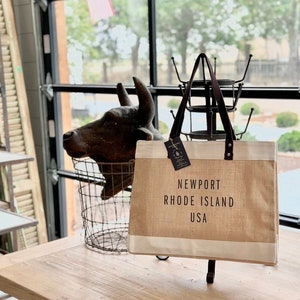 Op de afbeelding: Een bruine en witte tote bag met de tekst "NEWPORT RHODE ISLAND USA" erop gedrukt. De tas staat op een houten tafel voor een raam.