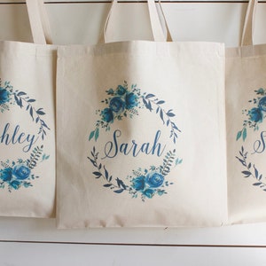 May include: Three white canvas tote bags with blue floral wreaths and the names "Ashley", "Sarah", and "Scarlett" printed on them.
