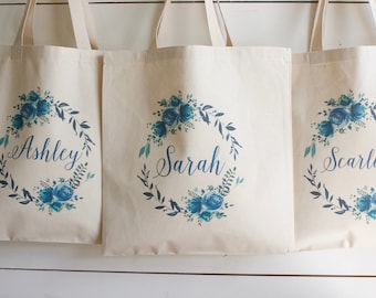 May include: Three white canvas tote bags with blue floral wreaths and the names "Ashley", "Sarah", and "Scarlett" printed on them.