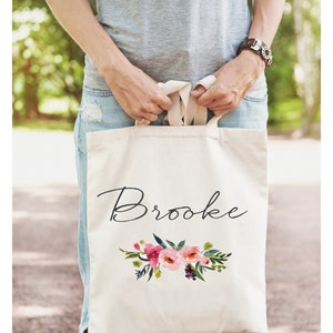 May include: A white canvas tote bag with the name "Brooke" written in black cursive script. The bag features a floral design with pink, red, and green watercolor flowers.