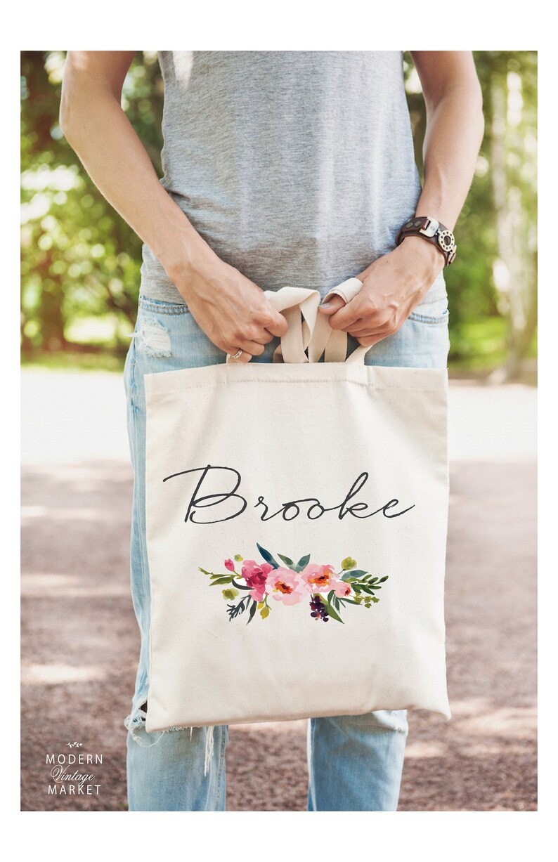 May include: A white canvas tote bag with the name "Brooke" written in black cursive script. The bag features a floral design with pink, red, and green watercolor flowers.