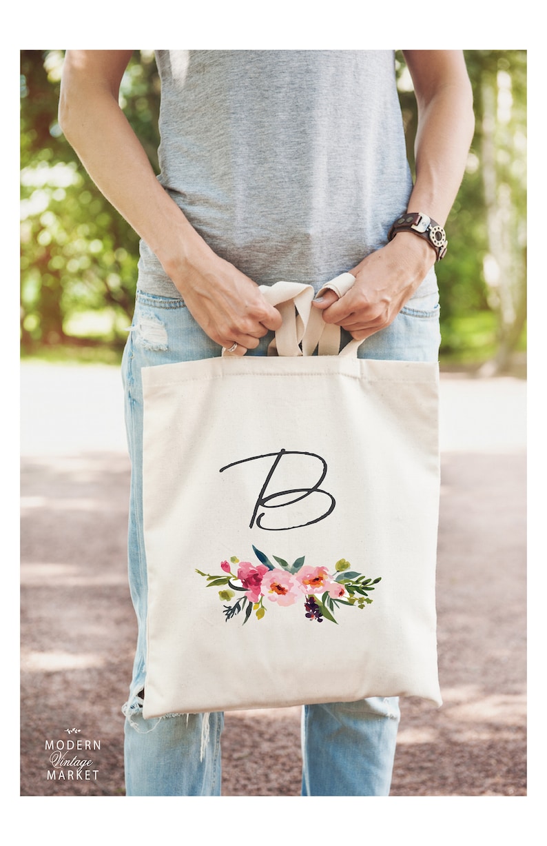 May include: A white canvas tote bag with a floral design and the letter "B" in black script. The bag has a pink, white, and green floral design with a dark green stem.