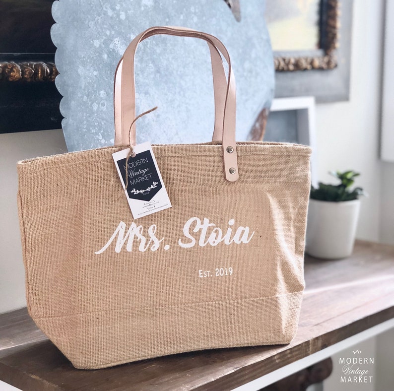 Op de afbeelding: Een natuurlijke jute tas met leren handvatten en een witte inscriptie die "Mrs. Stoia Est. 2019" luidt. De tas heeft een label met de tekst "Modern Vintage Market".