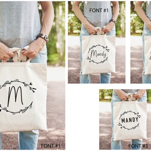 May include: Three tote bags with different fonts and designs. The first bag has a simple 'M' in a wreath. The second bag has the name 'Mandy' in a cursive font in a wreath. The third bag has the name 'MANDY' in a block font in a wreath.
