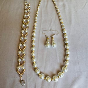 May include: A jewelry set featuring a pearl necklace, bracelet, and earrings. The necklace and bracelet have white pearls with gold-colored accents. The earrings are a drop style with three pearls each. The jewelry is displayed on a light purple surface.