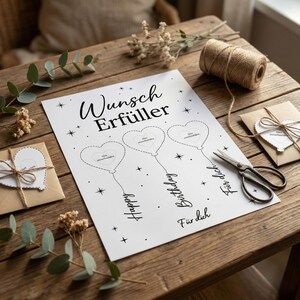 May include: A white paper with black text and heart-shaped balloon designs. The text reads "Wunsch Erf&uuml;ller", "Happy", "Birthday", and "F&uuml;r dich". Scissors, twine, and small envelopes are on a wooden surface.