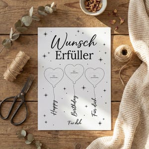 May include: A white card with black text and illustrations. The card reads "Wunsch Erf&uuml;ller" and features three heart-shaped balloons with the words "Happy", "Birthday", and "F&uuml;r dich" below them. The card is on a wooden surface.
