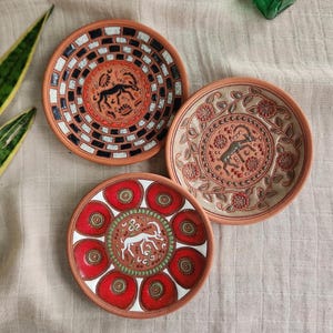 May include: Three round ceramic plates featuring hand-painted designs. One plate has a black and white checkered border with a central deer, another displays floral vines and a deer, and the third shows red circular motifs with a white deer.