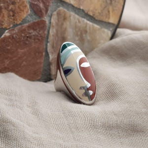 May include: A silver ring with an oval face design. The face is divided into sections of cream, red, and blue, with silver accents. The ring has a polished silver band and a unique, artistic appearance.