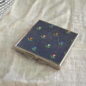 May include: A small, square, silver-toned compact with a navy blue top. The top is decorated with a repeating pattern of small flowers in orange, green, and blue. The compact has a hinged opening and a clasp.