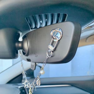 May include: A black car rearview mirror with a decorative silver chain hanging from it. The chain features a pearl and various charms. The mirror is mounted on the car's windscreen.