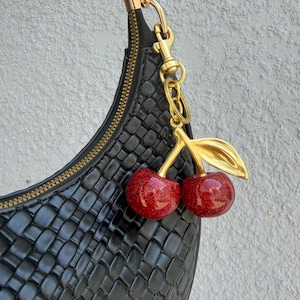 May include: Black woven handbag with a gold zipper and hardware. A gold cherry charm with red glitter cherries hangs from the strap. The bag has a crescent shape.