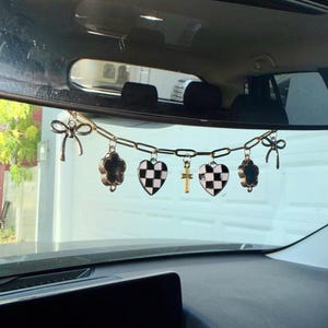 Puede incluir: Una cadena de metal dorado colgada de un espejo retrovisor, con seis dijes: dos lazos plateados en los extremos, dos corazones a cuadros blancos y negros, una pequeña cruz dorada y dos dijes en forma de flor negra con bordes plateados.
