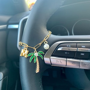 May include: Gold-toned car charm featuring a chain with a letter "M" charm, a green and gold palm tree charm with a clear gem, and a white pearl. The charm is attached to a black steering wheel.