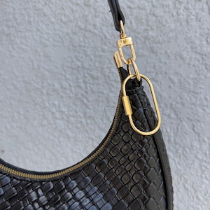 May include: Black woven leather shoulder bag with a gold-colored zipper and hardware. The bag has a curved shape and a single shoulder strap. The bag is hanging against a light gray textured wall.