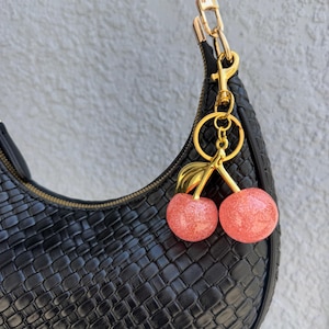 May include: Black woven handbag with a gold zipper and strap. A gold keychain with two glittery pink cherries hangs from the bag. The cherries have gold stems and leaves.