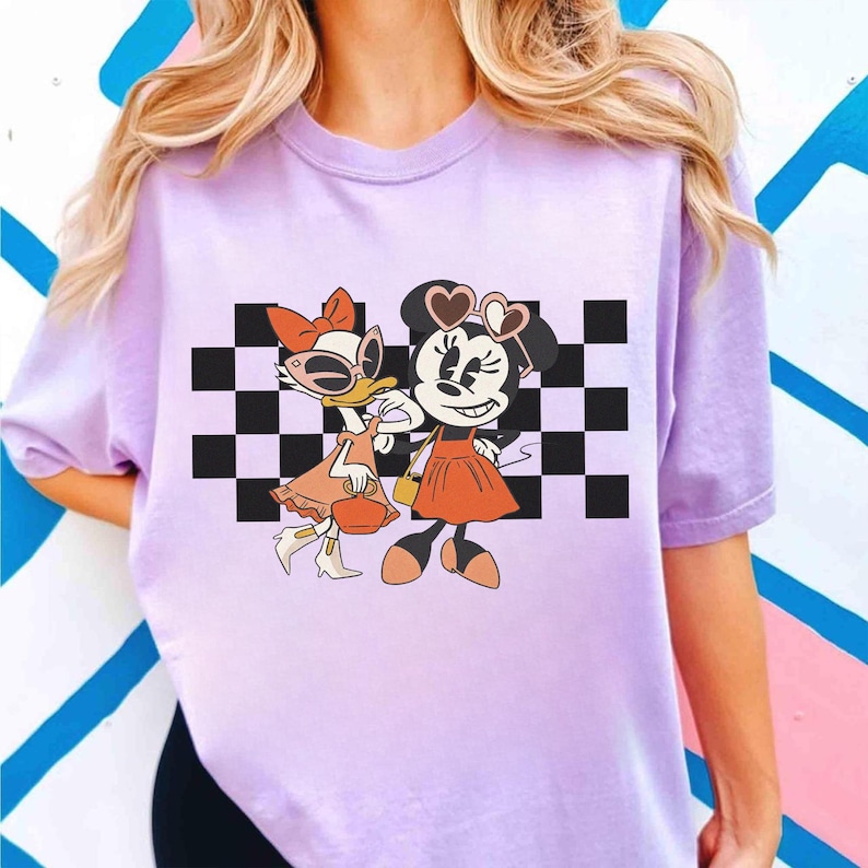 May include: A light purple t-shirt featuring a graphic of Daisy Duck and Minnie Mouse. The cartoon characters are wearing sunglasses and dresses, standing in front of a black and white checkered background.