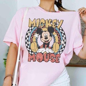 Puede incluir: Camiseta rosa claro con un dise&ntilde;o de Mickey Mouse. El dise&ntilde;o incluye las palabras "MICKEY" y "MOUSE" en una fuente retro, con Mickey en el centro, rodeado de un arco&iacute;ris y estrellas. Un patr&oacute;n de cuadros enmarca el dise&ntilde;o.