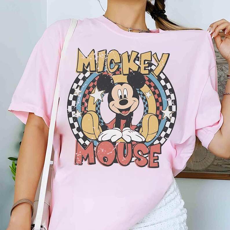 Puede incluir: Camiseta rosa claro con un dise&ntilde;o de Mickey Mouse. El dise&ntilde;o incluye las palabras "MICKEY" y "MOUSE" en una fuente retro, con Mickey en el centro, rodeado de un arco&iacute;ris y estrellas. Un patr&oacute;n de cuadros enmarca el dise&ntilde;o.