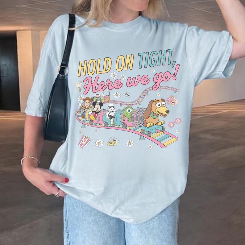 May include: Light blue t-shirt featuring a colorful graphic of a toy train with Disney characters and the text "HOLD ON TIGHT, Here we go!". The shirt has short sleeves and a relaxed fit.