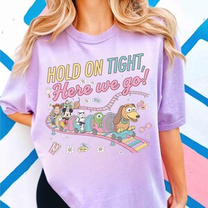 May include: A light purple t-shirt with the phrase "HOLD ON TIGHT, Here we go!" in yellow and pink lettering. The shirt features a cartoon roller coaster with characters like Woody, Mickey Mouse, and Slinky Dog.