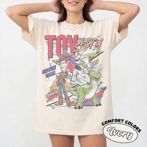 Puede incluir: Camiseta de color marfil con un gr&aacute;fico de los personajes de Toy Story Woody, Buzz Lightyear y Rex. El dise&ntilde;o incluye el texto "Toy Story" en rosa, "Since 1995" y "Woody". La camiseta tambi&eacute;n tiene las palabras "Comfort Colors" e "Ivory".