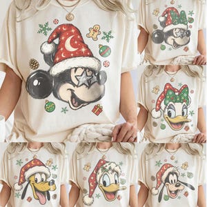 Könnte beinhalten: Ein cremefarbenes T-Shirt mit einer Grafik einer Cartoon-Maus, die eine rote Weihnachtsmütze mit Mond- und Sternenmustern sowie sternförmige Brillen trägt, umgeben von kleinen festlichen Symbolen wie Lebkuchenmännern, Tannenzapfen und Kugeln.