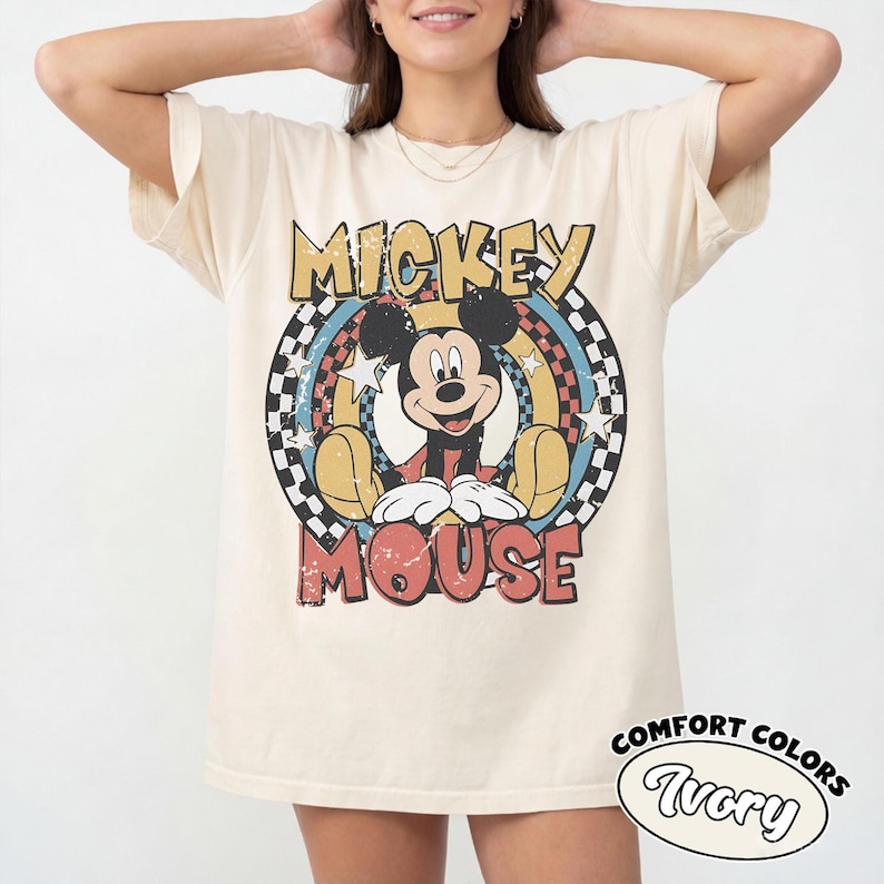 Puede incluir: Camiseta de color marfil con un gr&aacute;fico desgastado de Mickey Mouse. El dise&ntilde;o incluye las palabras "Mickey Mouse" en una fuente retro, rodeadas por un dise&ntilde;o circular colorido con estrellas y un patr&oacute;n de cuadros. La camiseta tambi&eacute;n tiene las palabras "Comfort Colors" e "Ivory".
