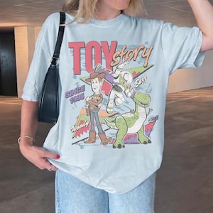 Puede incluir: Camiseta azul claro con un dise&ntilde;o de Woody, Buzz Lightyear y Rex de Toy Story. El dise&ntilde;o incluye el texto "Toy Story" y "Since 1995" en un estilo retro. Una prenda casual.