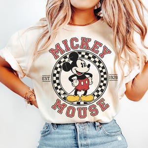 May include: Cream-colored t-shirt featuring a vintage-style Mickey Mouse graphic. The design includes the words "Mickey Mouse" in red, with "EST. 1928" and a checkered circle. The cartoon character is in red shorts and yellow shoes.