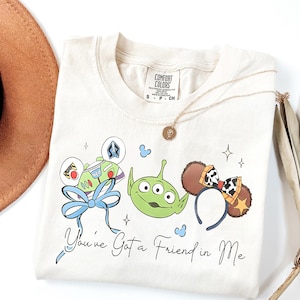 以下が含まれることがあります： クリーム色のTシャツ。Buzz Lightyear、緑色のエイリアン、ミッキーマウスの耳の漫画のキャラクターと「You've Got a Friend in Me」のフレーズが特徴です。金のネックレスと茶色の帽子も表示されています。