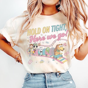 Può includere: T-shirt color crema con la scritta "HOLD ON TIGHT, Here we go!" in giallo e rosa. La grafica presenta personaggi dei cartoni animati sulle montagne russe. Il design include Woody, Mickey Mouse, uno Stormtrooper e Slinky Dog.