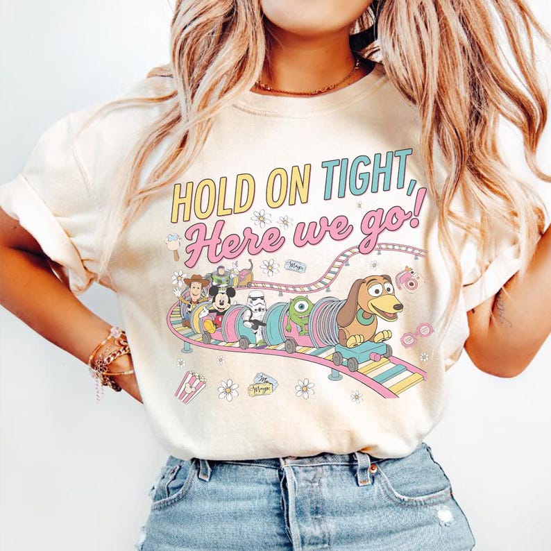 May include: Cream-colored t-shirt with the text "HOLD ON TIGHT, Here we go!" in yellow and pink. The graphic features cartoon characters on a roller coaster. The design includes Woody, Mickey Mouse, a Stormtrooper, and Slinky Dog.